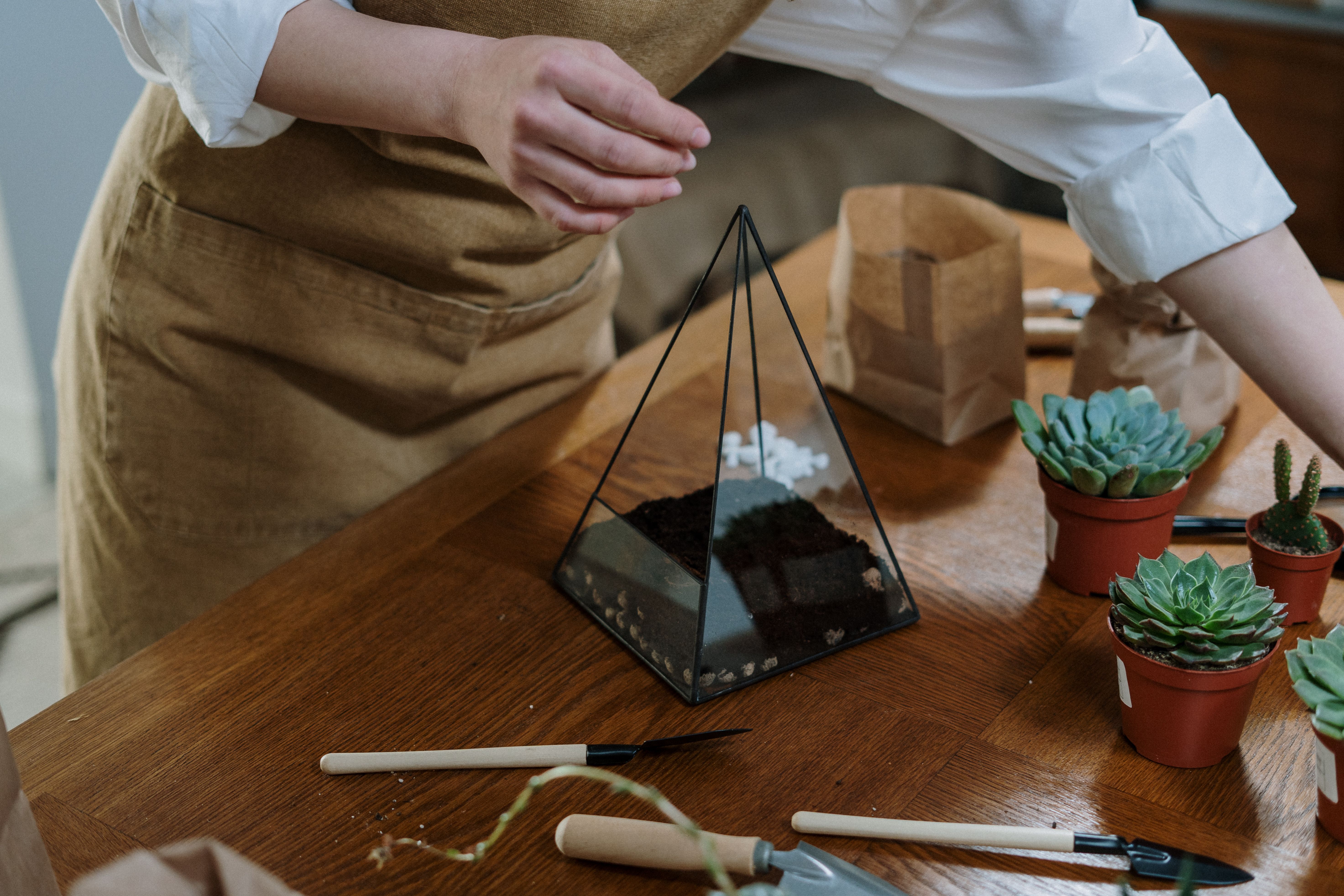 Mãos montando um terrário com camadas de terra e pedras em recipiente de vidro