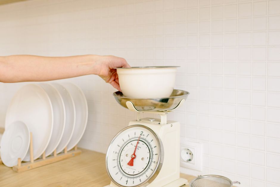 Mão colocando tigela sobre balança de cozinha digital para zerar o peso do recipiente