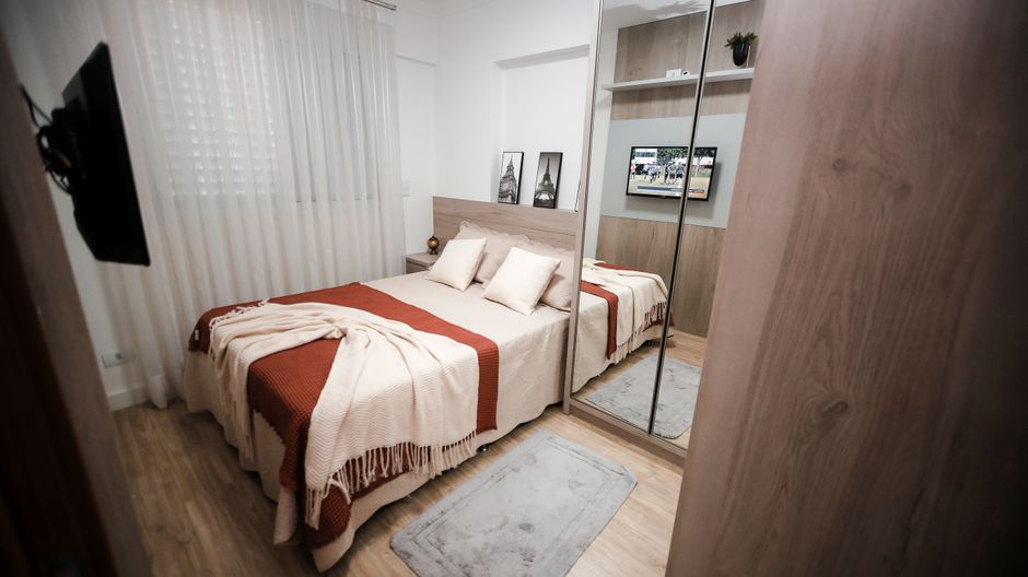 Quarto de casal compacto com cama ocupando quase toda a largura e guarda-roupa espelhado ao lado