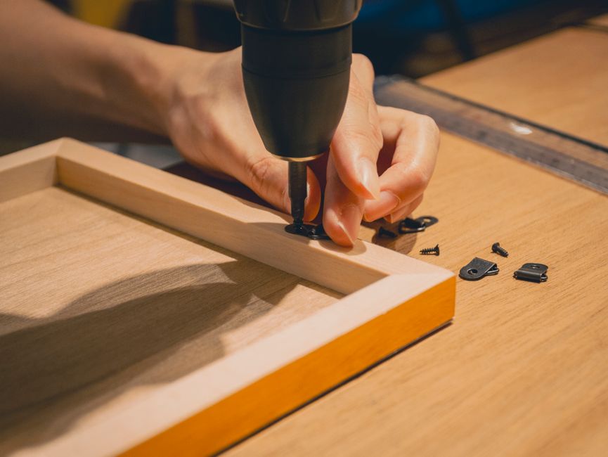 Mãos usando furadeira para fixar sarrafo de madeira em moldura durante projeto DIY